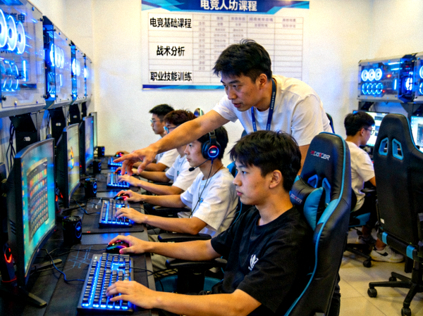 爱游戏电竞人才培养基地实景：学员在专业设备前进行训练，教练在一旁指导，背景是各类电竞课程安排表
